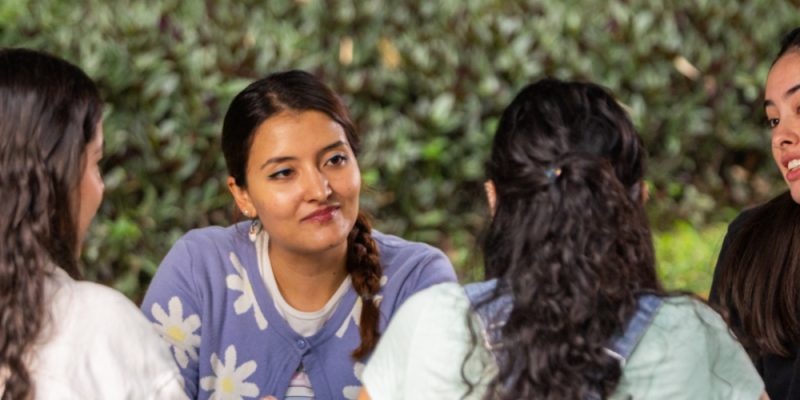 Cuatro personas sentadas en círculo conversando al aire libre, con vegetación en el fondo.