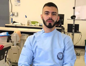 Persona con bata clínica azul está sentada junto a una silla odontológica en un consultorio equipado para prácticas dentales.