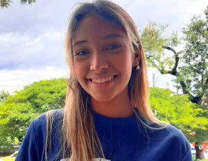 Persona con cabello largo y lacio, vestida con camiseta azul, está en un espacio al aire libre con árboles y cielo parcialmente nublado en el fondo.