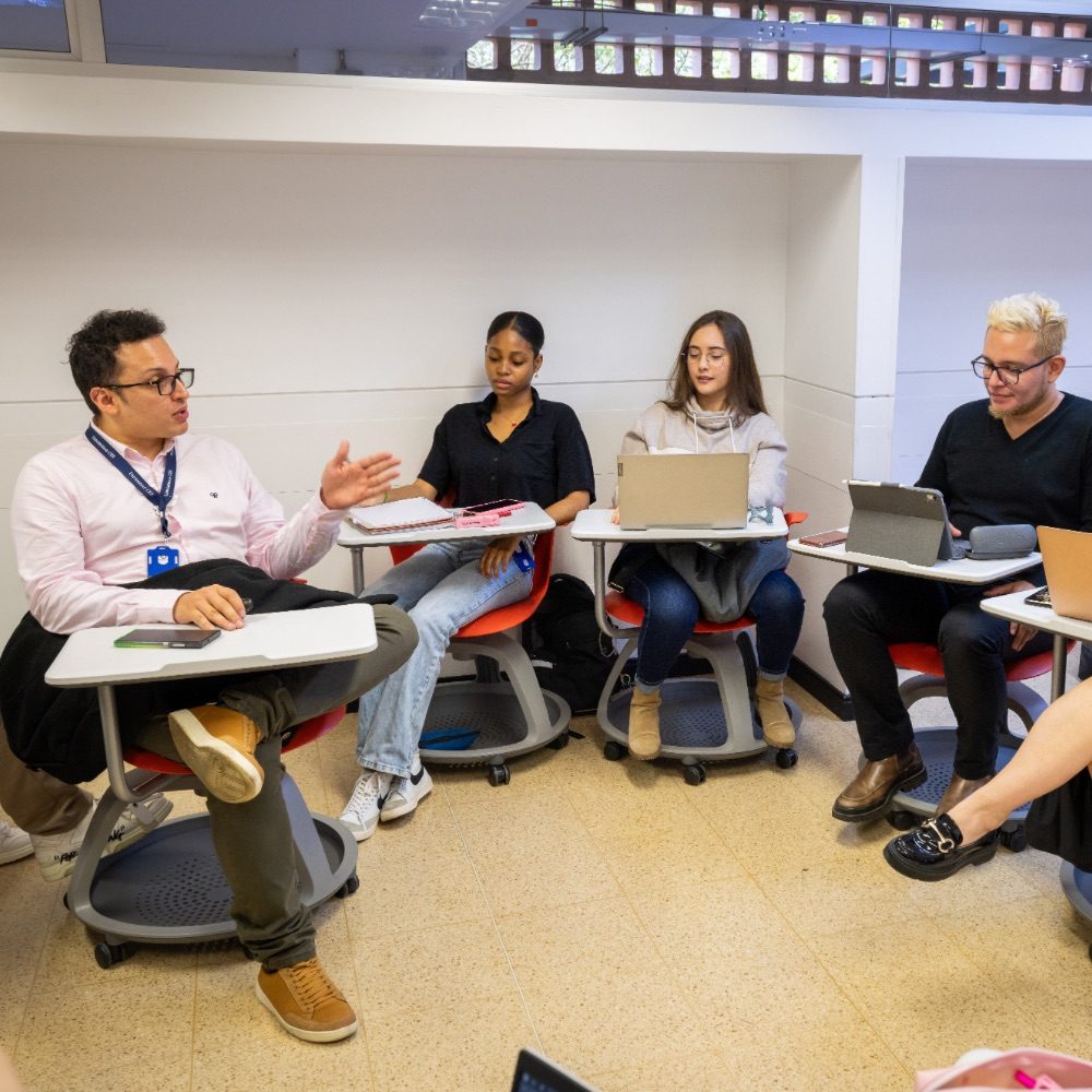 Varias personas están sentadas en un aula en disposición circular, utilizando portátiles y cuadernos mientras participan en una conversación grupal.