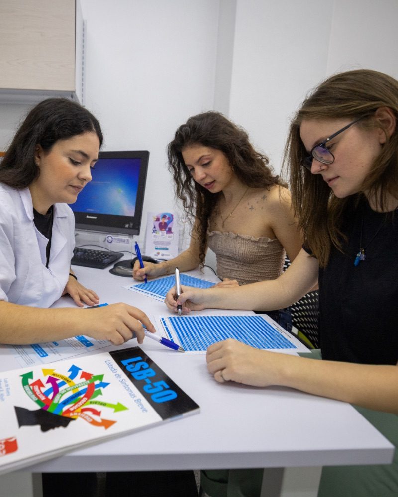 Tres personas están sentadas alrededor de una mesa realizando una actividad con hojas impresas y bolígrafos. Sobre la mesa hay un cuaderno con un diseño colorido y una computadora en el fondo.