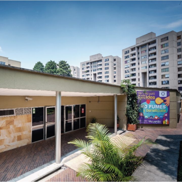 Entrada de un edificio con paredes claras y puertas de vidrio, junto a un cartel colorido con mensaje preventivo y edificios residenciales al fondo.