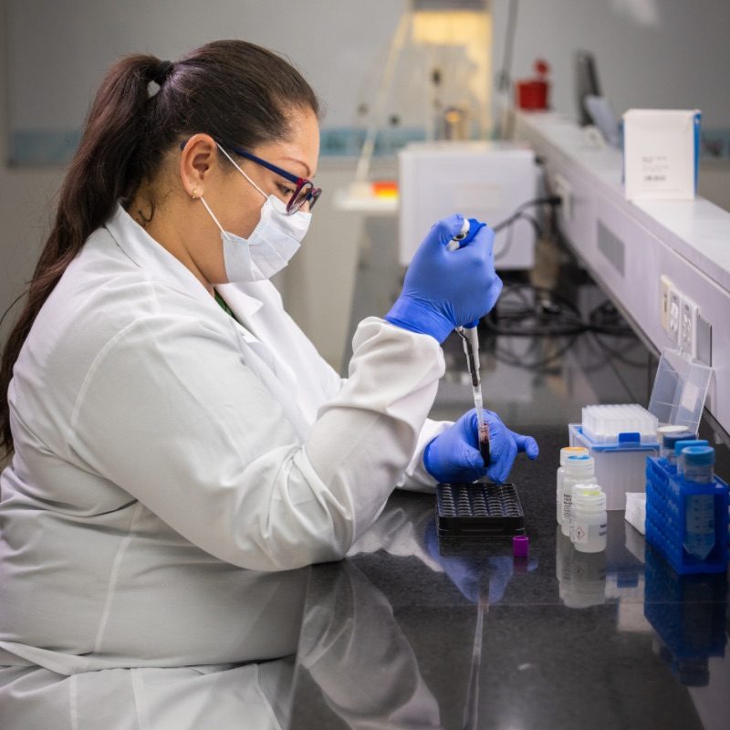 Persona sentada en un escritorio frente a un computador y un cartel de la tabla periódica en la pared.