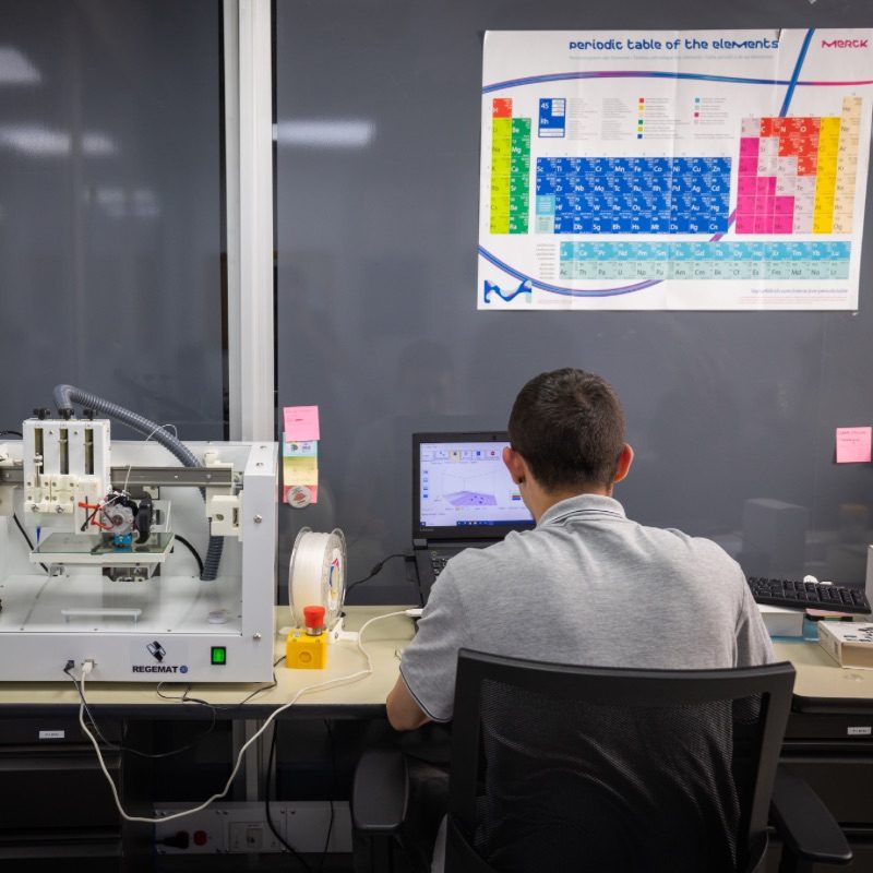 Persona sentada en un escritorio frente a un computador, con una impresora 3D a un lado y un cartel de la tabla periódica en la pared.