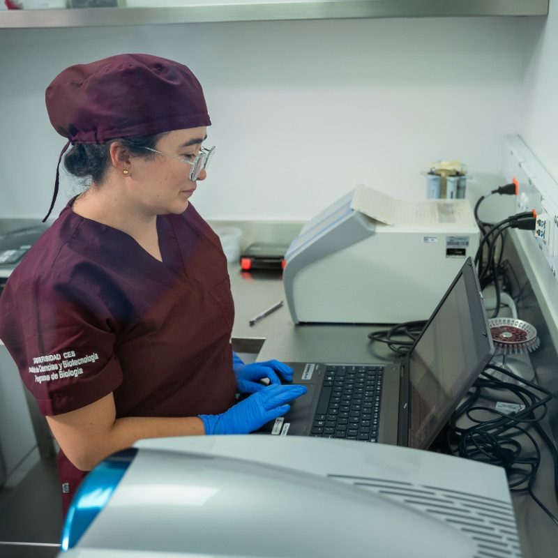 Persona con uniforme de laboratorio y guantes azules trabajando en un computador portátil sobre una mesa, junto a equipos de laboratorio.
