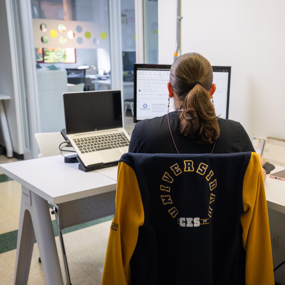 Persona sentada frente a un escritorio con un computador portátil y un monitor encendido. Sobre la silla cuelga una chaqueta con el texto “Universidad CES”. Al fondo se observa una oficina con paredes blancas y un ventanal con círculos de colores.