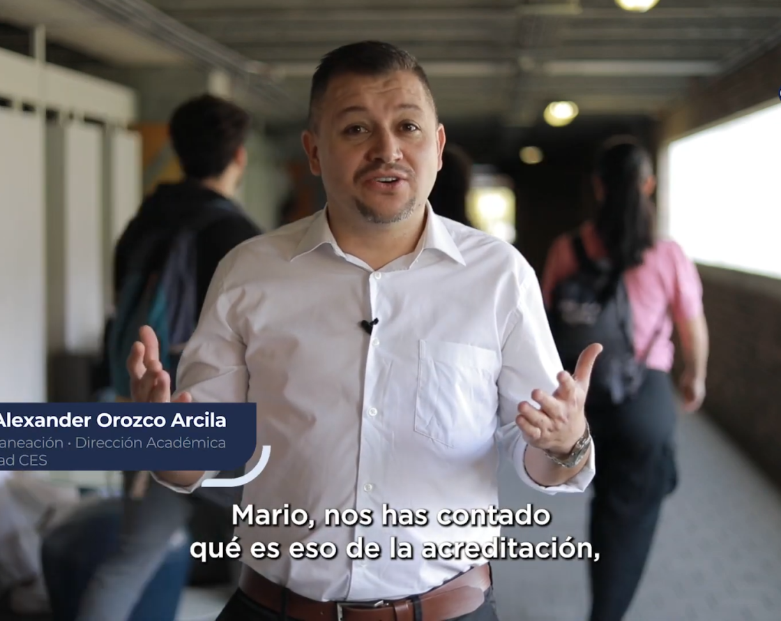 Persona de pie en un pasillo interior, con otras personas al fondo, sosteniendo un micrófono de solapa y texto en pantalla sobre acreditación institucional.