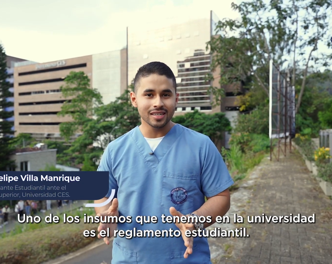 Persona con uniforme azul habla frente a la cámara en un sendero al aire libre, con edificios y vegetación en el fondo, y texto en pantalla sobre reglamento estudiantil.