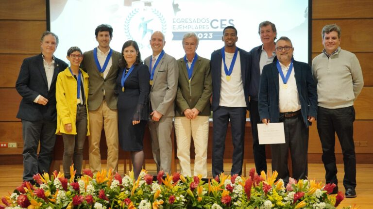 Grupo de personas posando en un escenario frente a un arreglo floral, con medallas colgadas al cuello y una pantalla de presentación en el fondo.