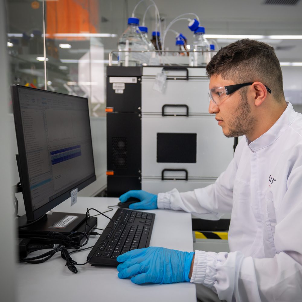 Persona con bata blanca y guantes azules trabajando en un computador dentro de un laboratorio, con equipos científicos al fondo.