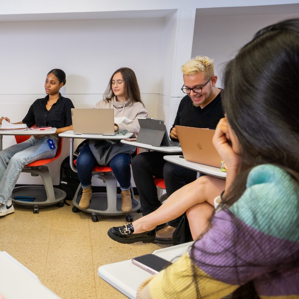 Varias personas sentadas en un aula, usando portátiles y cuadernos, organizadas en escritorios individuales.