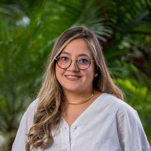 Persona de pie en un entorno exterior con vegetación verde, usando blusa blanca de manga larga con escote en V y un collar dorado sencillo.
