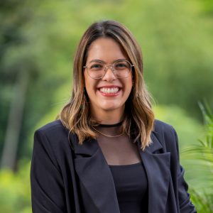 Persona de pie en un entorno exterior con vegetación verde, usando chaqueta negra sobre blusa negra con detalles transparentes en la parte superior y un collar dorado sencillo.