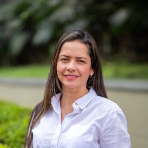 Persona de pie en un entorno exterior con vegetación desenfocada al fondo, usando camisa blanca de manga larga con cuello y botones visibles.