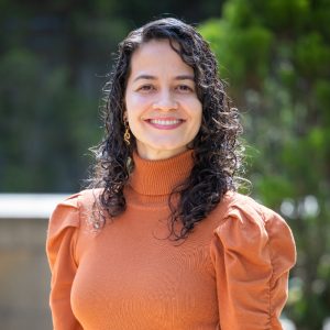 Persona con cabello rizado y largo, vistiendo suéter de cuello alto color naranja con mangas abullonadas, en un entorno exterior con vegetación desenfocada al fondo.