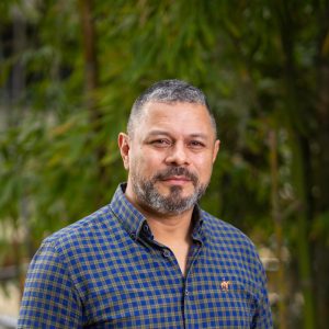 Persona con cabello corto y canoso, vistiendo camisa de cuadros azul y verde con cuello y botones, en un entorno exterior con vegetación desenfocada al fondo.