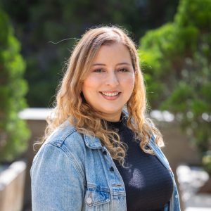 Persona con cabello largo y ondulado, vistiendo camiseta negra y chaqueta de mezclilla azul, en un entorno exterior con vegetación desenfocada al fondo.
