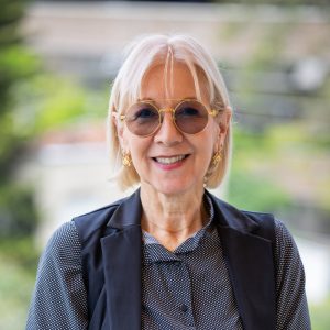 Persona con cabello claro y corto, usando blusa oscura con puntos y chaleco negro, en un entorno exterior con vegetación desenfocada al fondo.