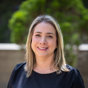 Persona con cabello lacio y rubio oscuro, usando blusa negra de cuello redondo, en un entorno exterior con vegetación desenfocada al fondo.