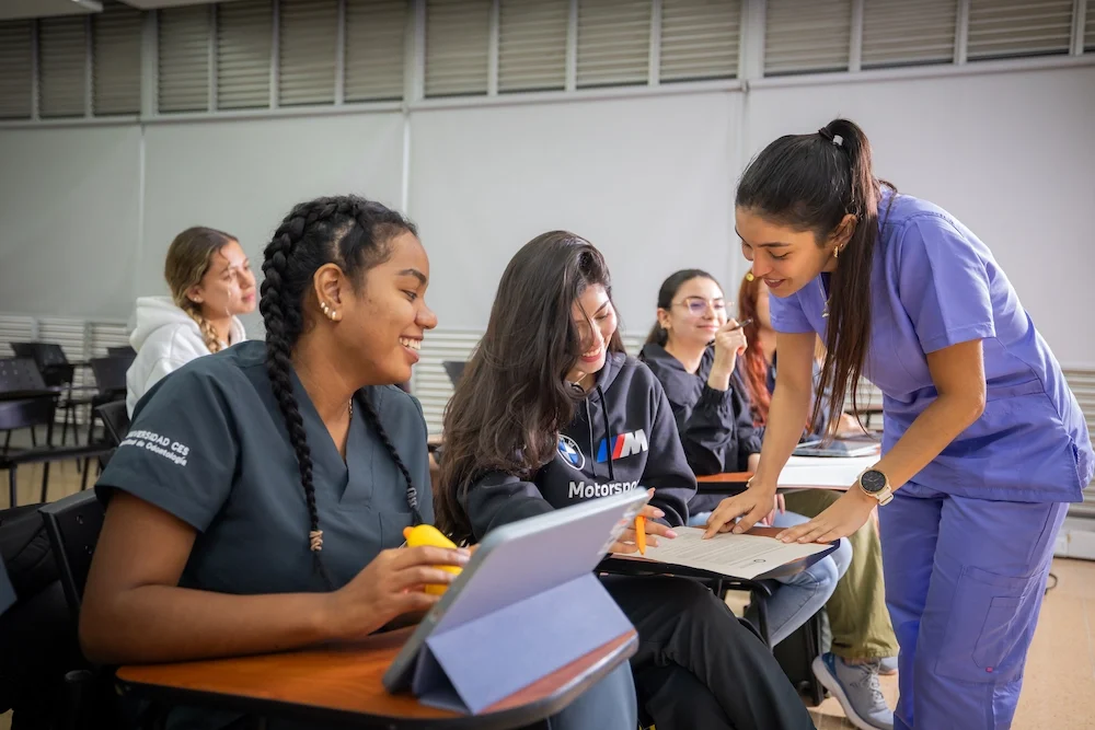 Varias personas están en un aula, algunas sentadas frente a escritorios con cuadernos y dispositivos, mientras otra persona de pie parece explicar una actividad. El entorno incluye sillas, mesas y una pared con ventanas en la parte superior.