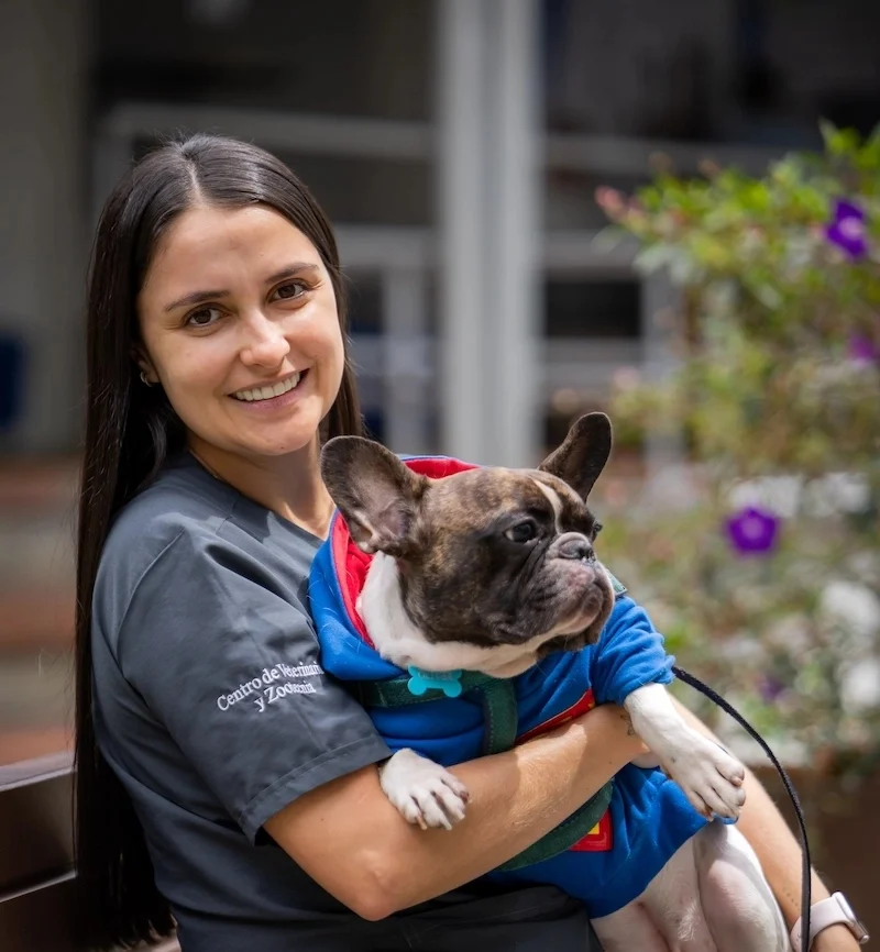 Estudiante sostiene en brazos a un perro de raza bulldog francés, que lleva puesta una chaqueta azul con detalles rojos. Al fondo se observan plantas con flores moradas y un entorno exterior.