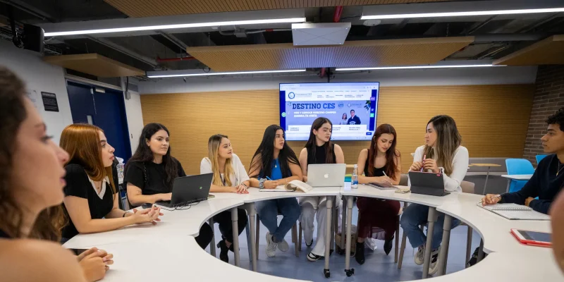 Grupo de personas reunidas alrededor de una mesa circular en un aula moderna; varias usan computadores portátiles y al fondo hay una pantalla con contenido proyectado.