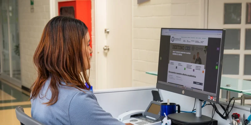 Persona sentada frente a un escritorio utiliza un computador de escritorio que muestra una página web institucional, en un espacio interior con paredes claras y objetos de oficina alrededor.